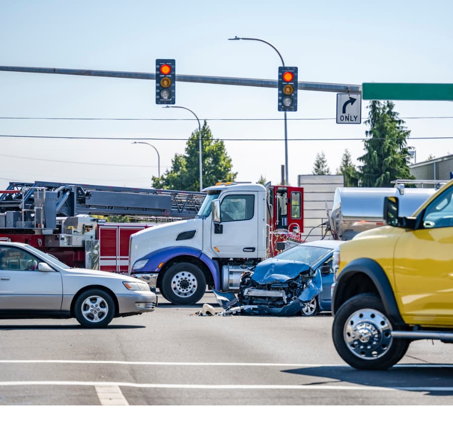 A crashed blue car. tanker truck, and fire engine in the middle of an intersection with a yellow truck and white car in the foreground