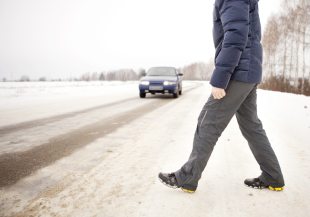 A person in a blue jacket and gray pants crossing a snowy road with a car heading toward them