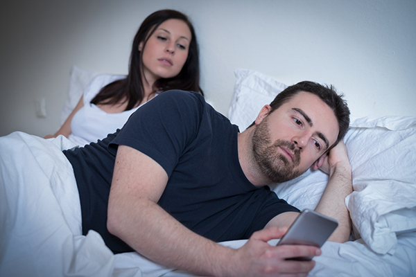 couple in bed, man is turned over looking at phone and woman is trying to see who he’s talking to