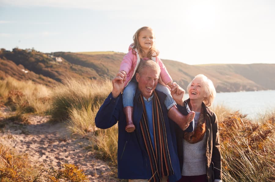 grandparents walking through sand dunes with granddaughter on grandfather’s shoulders