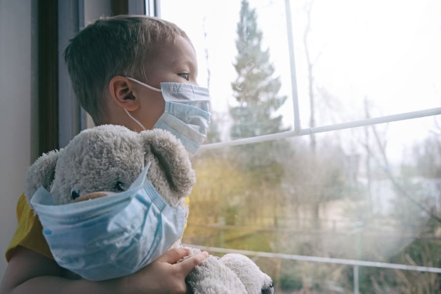 little boy holding his teddy bear looking out the window, both wearing a protective medical mask
