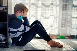 an upset young boy sitting on the floor wearing a protective medical mask, holding his head in both hands