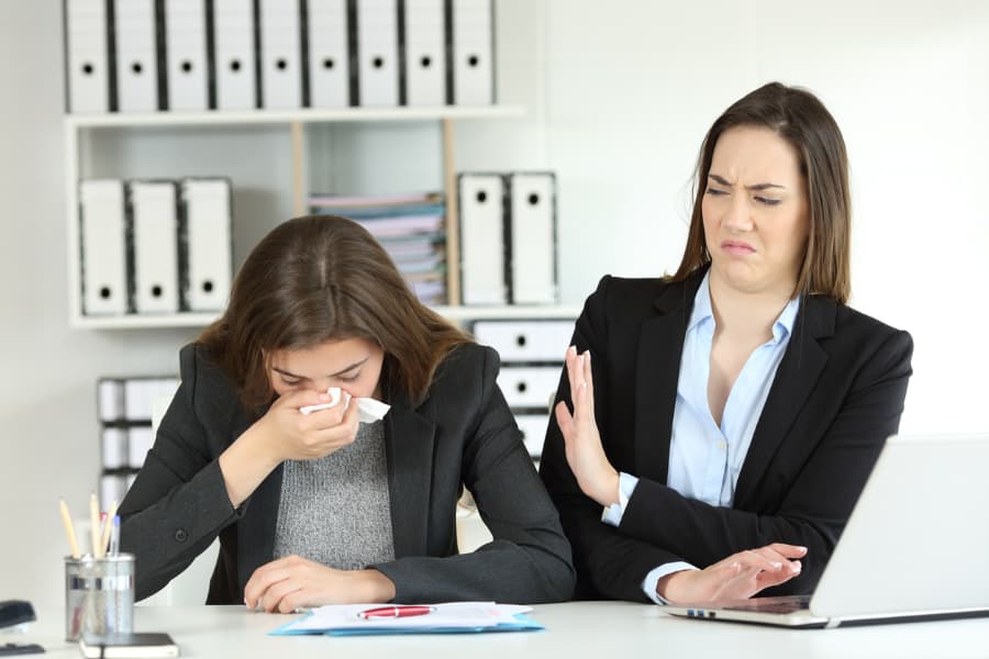 2 businesswomen standing at a desk, one coughing into a tissue and the other leaning away with her left hand up as a shield