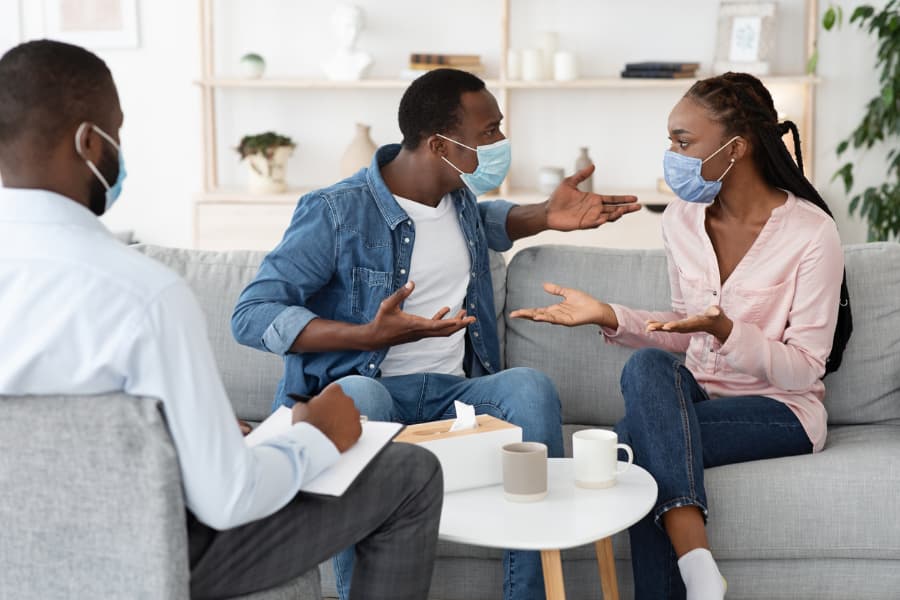 Young couple wearing masks arguing while sitting on couch in therapist’s office