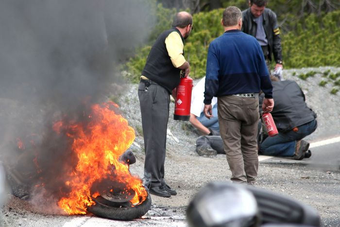 a traffic accident scene where a motorcycle is on fire and the victim is being attended to by civilians