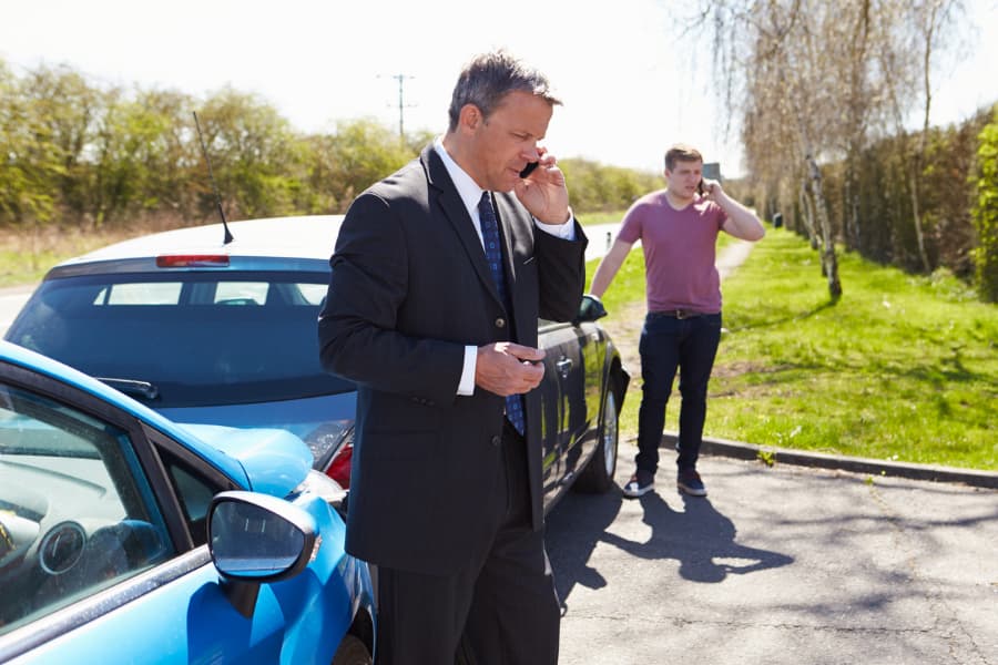 a businessman and young man standing next to a car accident talking on phones