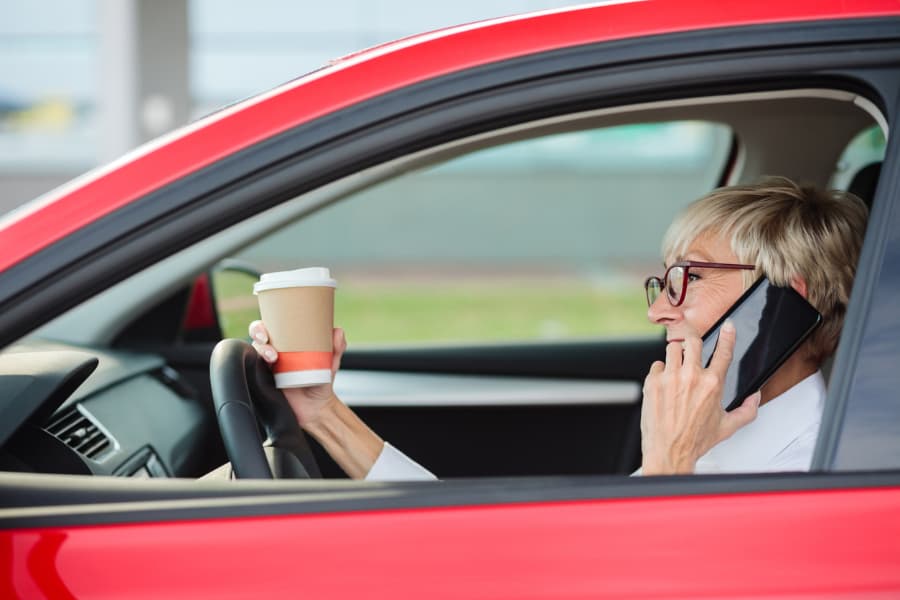 older woman driving car while talking on phone and drinking coffee
