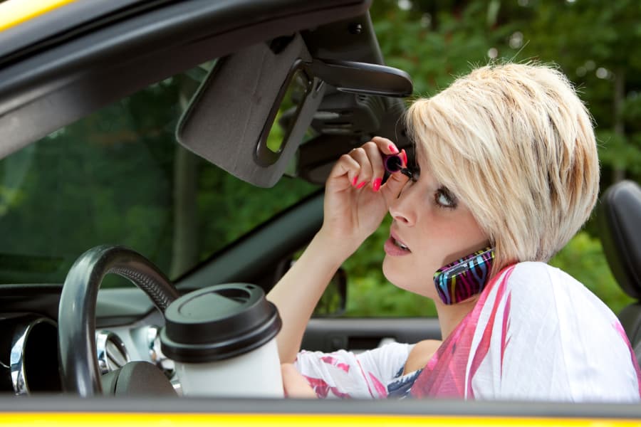 A woman applies make-up, talks on the phone, and drinks coffee while driving