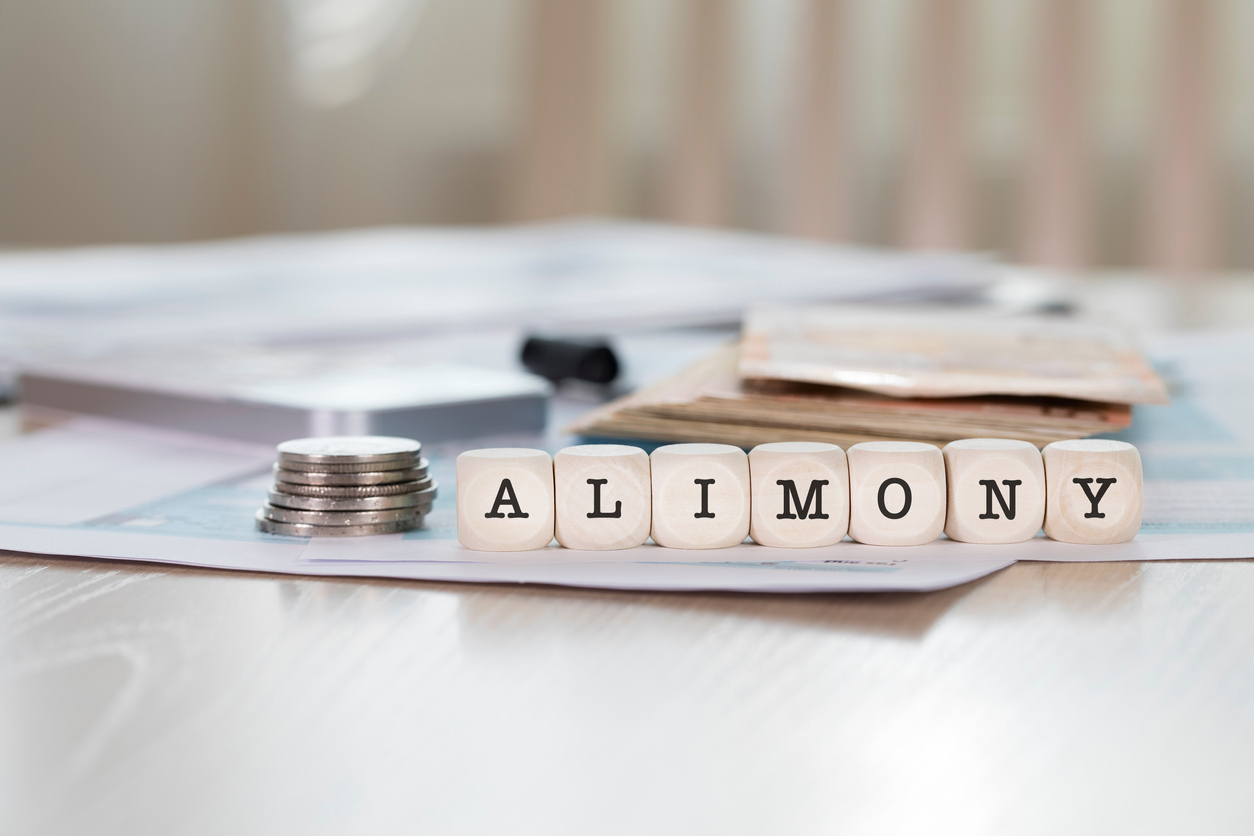 Close-up of the word “alimony” on small wooden blocks with a stack of coins and papers