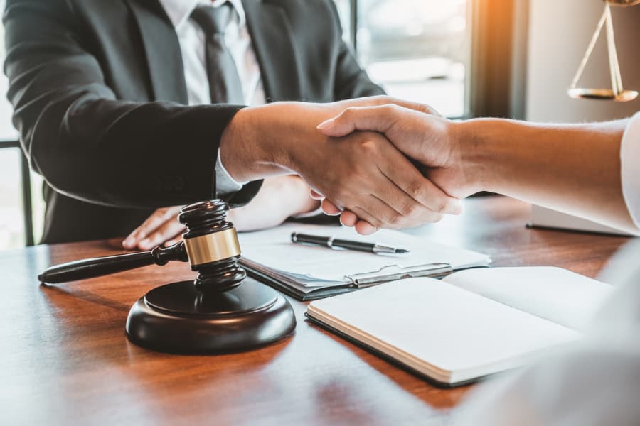 Close-up of a car accident lawyer and client shaking hands over a wooden desk with a gavel, notebook, clipboard and pen